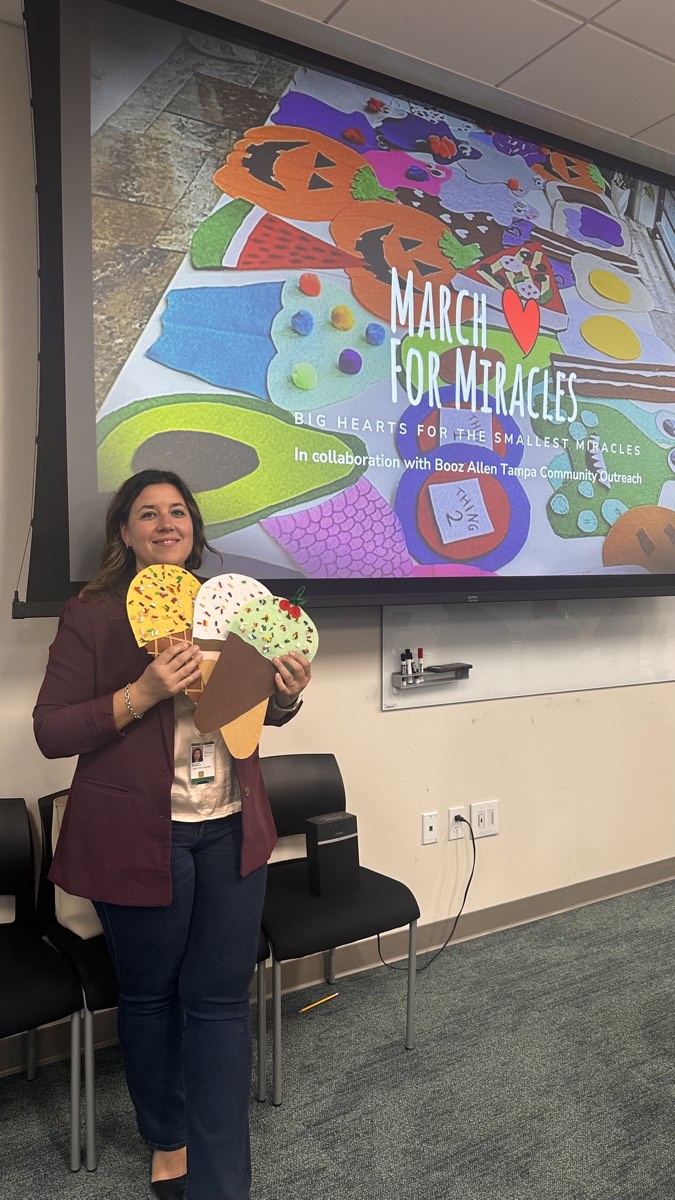 A volunteer holding colorful handmade costume pieces beside a March for Miracles presentation screen.