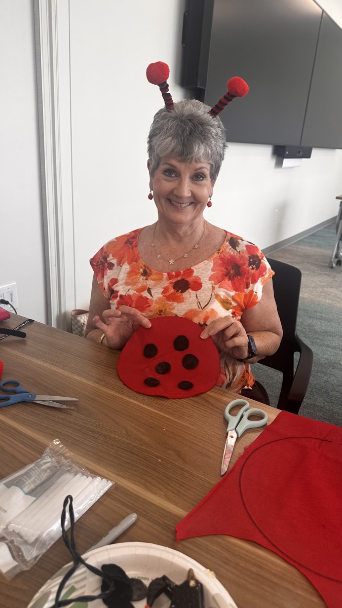 A smiling volunteer holding a handmade red and black ladybug costume at a craft table.